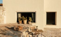 Modern minimalist house exterior with a stone terrace, featuring potted cacti and a small wooden table with chairs, under a clear blue sky.