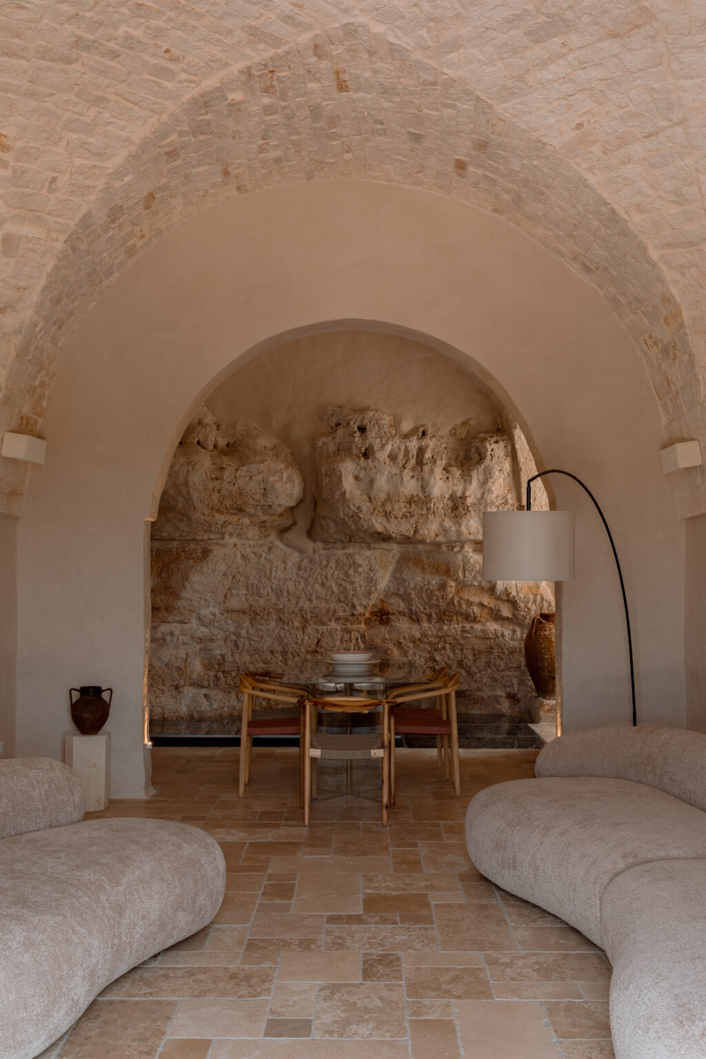 Modern interior design featuring a cozy seating area with curved sofas, a glass dining table, and a stylish floor lamp, set against a textured stone wall and arched ceiling. Ideal for showcasing contemporary home decor inspiration. | Only on SopranoVillas