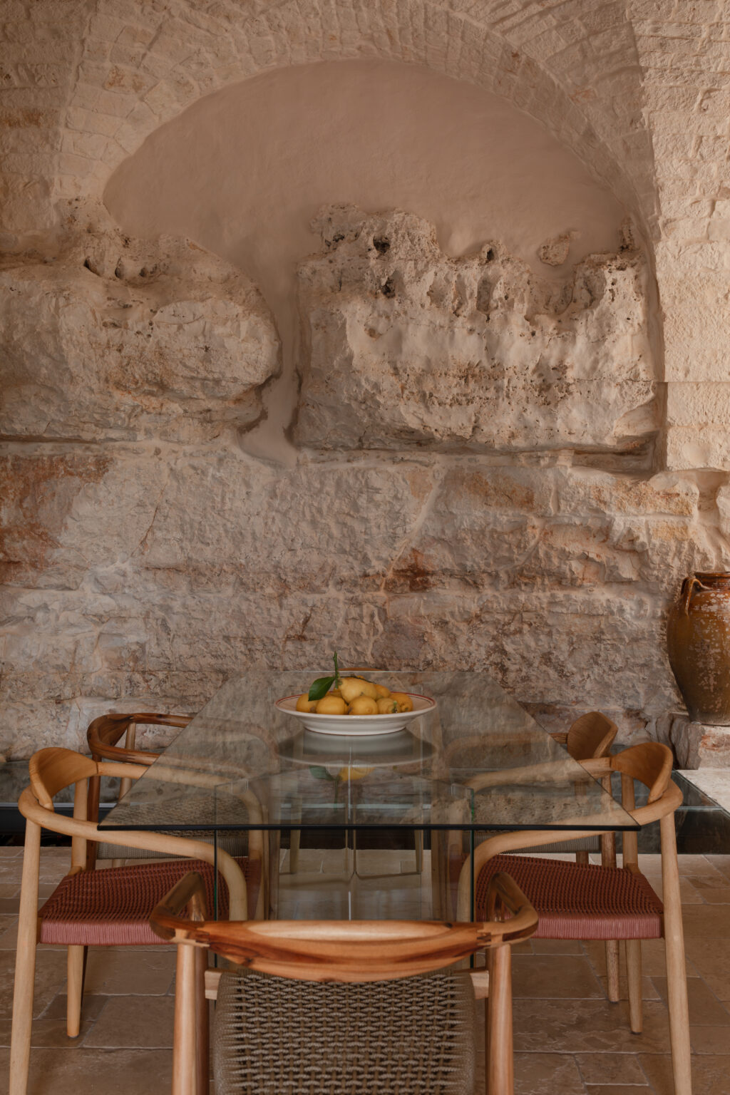Modern dining area featuring a glass table surrounded by wooden chairs, with a bowl of fresh lemons as a centerpiece, set against a rustic stone wall with an arched design. | Only on SopranoVillas