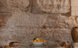 Modern dining area featuring a glass table surrounded by wooden chairs, with a bowl of fresh lemons as a centerpiece, set against a rustic stone wall with an arched design.