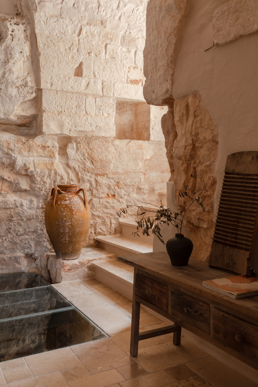 Interior view of a rustic space featuring a large terracotta vase, a wooden table with decorative items, and natural stone walls. A glass section in the floor reveals the underlying structure, and stairs lead to another level, enhancing the room's depth and character. | Only on SopranoVillas