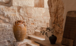 Interior view of a rustic space featuring a large terracotta vase, a wooden table with decorative items, and natural stone walls. A glass section in the floor reveals the underlying structure, and stairs lead to another level, enhancing the room's depth and character.