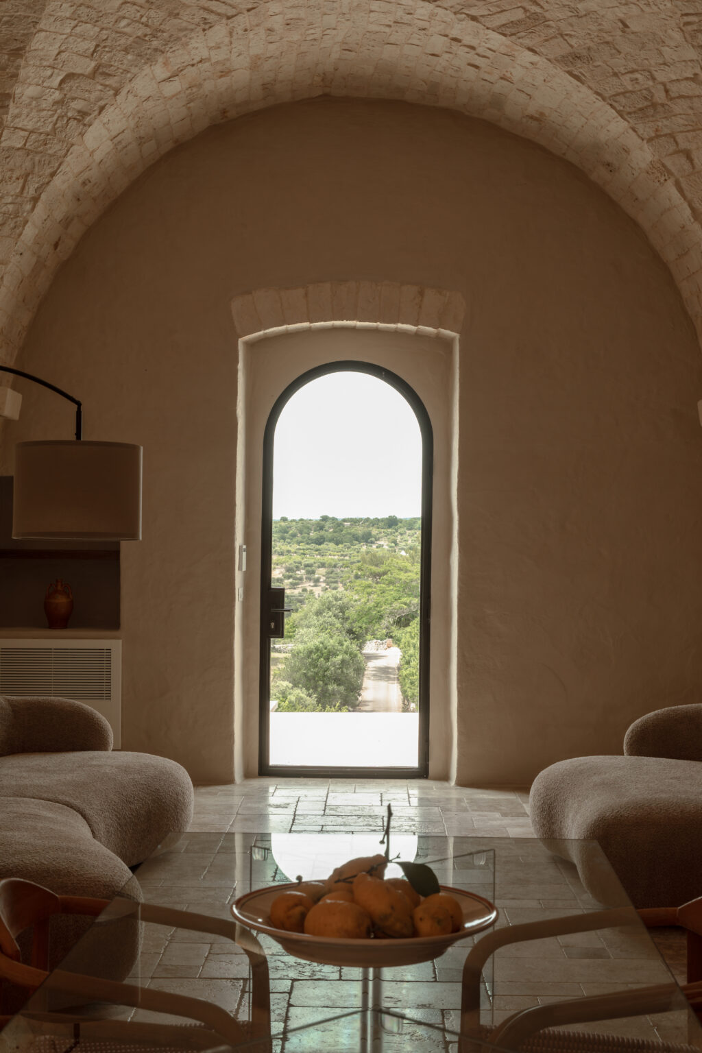 Interior view of a cozy living space featuring a large arched doorway leading to a lush outdoor landscape, with comfortable seating and a glass coffee table displaying a bowl of fresh lemons. The warm, earthy tones and natural light create a serene atmosphere. | Only on SopranoVillas