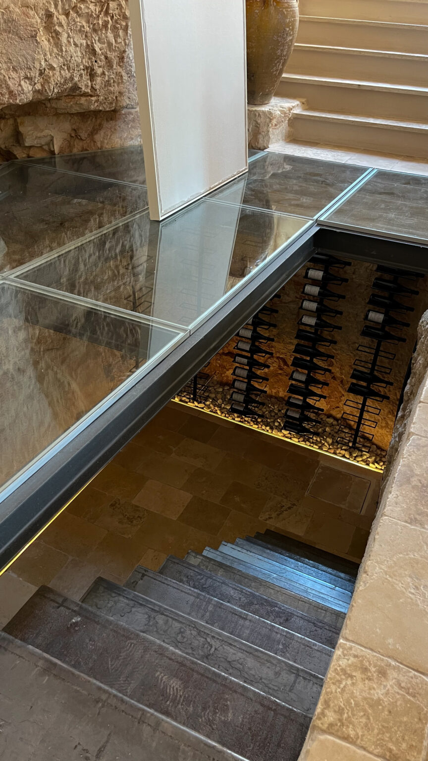 Modern staircase leading to a wine cellar, featuring glass flooring that showcases wine racks below, surrounded by stone walls and ambient lighting. | Only on SopranoVillas