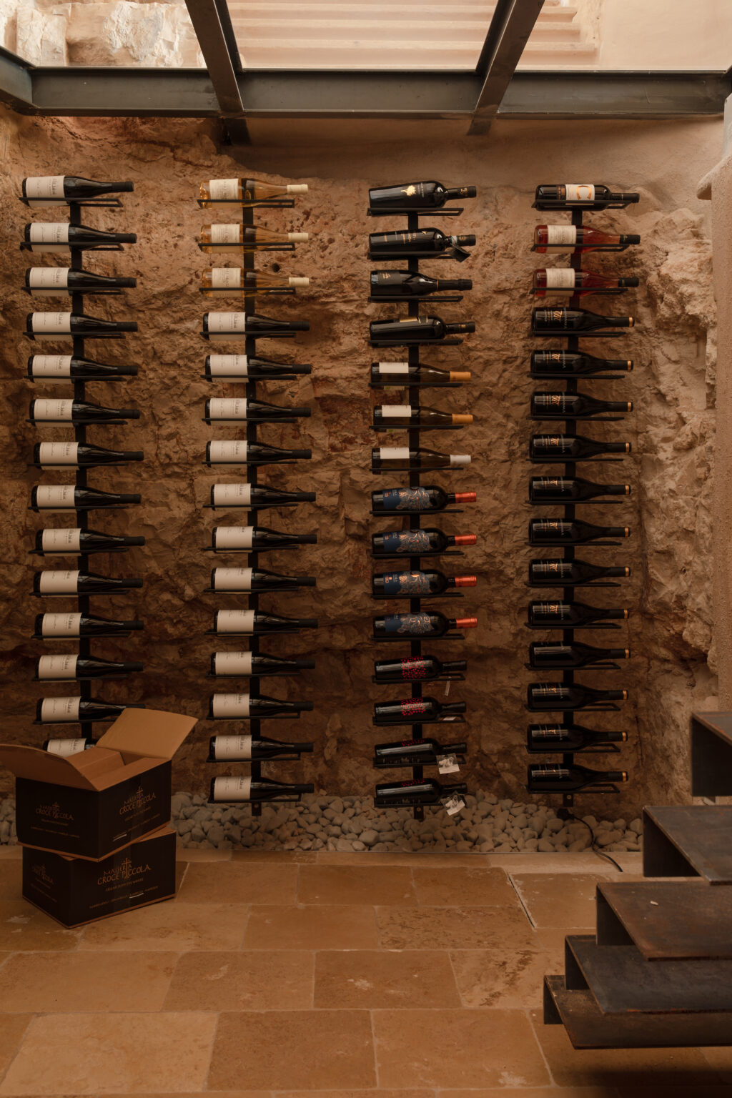 Wine cellar featuring neatly arranged wine bottles on wall-mounted racks against a natural stone backdrop, with a cardboard box on the floor and stone flooring. | Only on SopranoVillas