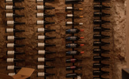 Wine cellar featuring neatly arranged wine bottles on wall-mounted racks against a natural stone backdrop, with a cardboard box on the floor and stone flooring.