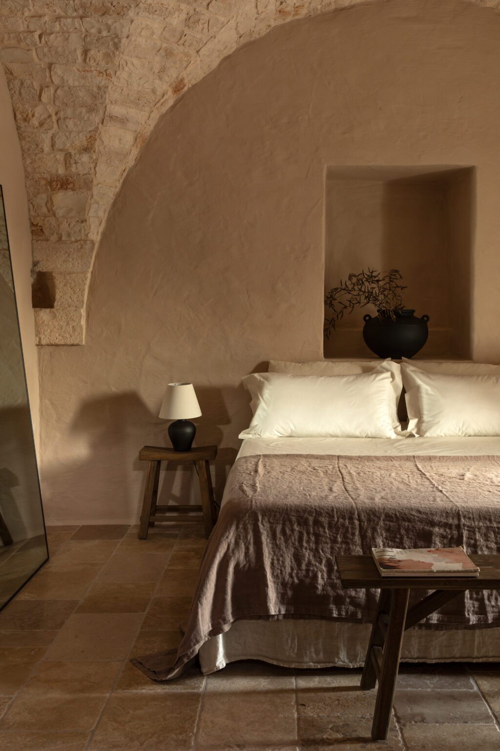 Cozy bedroom featuring a neutral color palette with a stone archway, a comfortable bed with white pillows, a textured brown blanket, a wooden side table with a lamp, and decorative elements including a plant and a black vase. The room's serene ambiance is enhanced by natural light and rustic decor. | Only on SopranoVillas