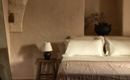 Cozy bedroom featuring a neutral color palette with a stone archway, a comfortable bed with white pillows, a textured brown blanket, a wooden side table with a lamp, and decorative elements including a plant and a black vase. The room's serene ambiance is enhanced by natural light and rustic decor.