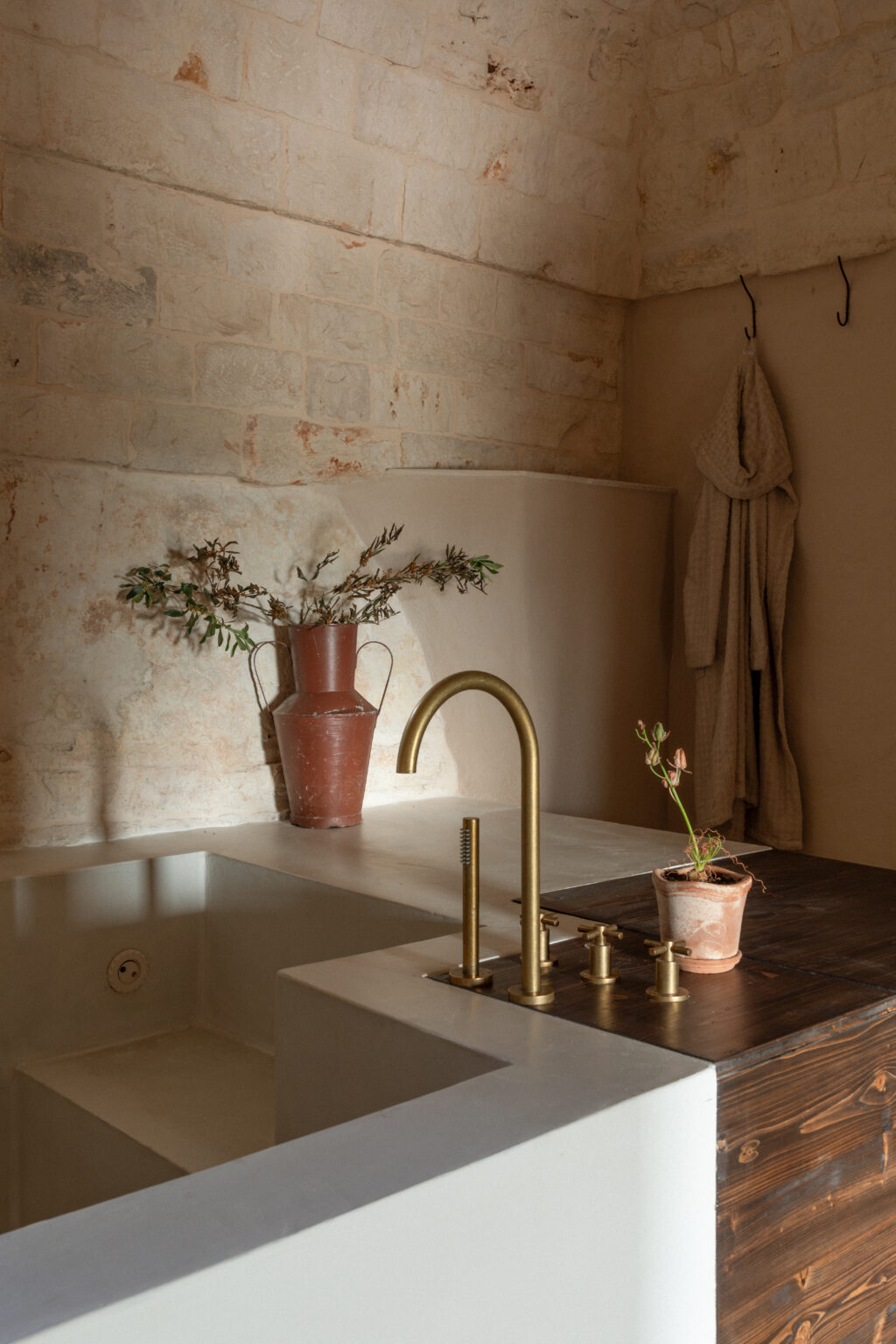 Modern bathroom interior featuring a minimalist design with a brass faucet, a rustic wooden countertop, and a terracotta vase holding dried foliage. The textured stone walls and soft lighting create a warm and inviting atmosphere. | Only on SopranoVillas