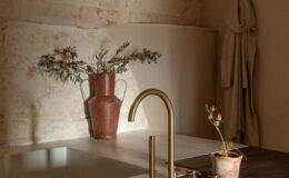 Modern bathroom interior featuring a minimalist design with a brass faucet, a rustic wooden countertop, and a terracotta vase holding dried foliage. The textured stone walls and soft lighting create a warm and inviting atmosphere.