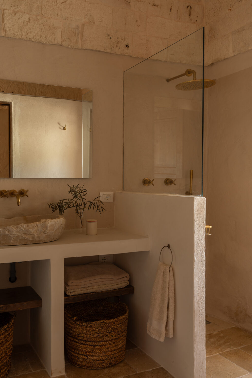 Modern bathroom interior featuring a minimalist design with natural stone walls, a glass shower enclosure, a sculptural sink, brass fixtures, and woven storage baskets, creating a serene and stylish atmosphere. | Only on SopranoVillas