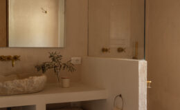 Modern bathroom interior featuring a minimalist design with natural stone walls, a glass shower enclosure, a sculptural sink, brass fixtures, and woven storage baskets, creating a serene and stylish atmosphere.