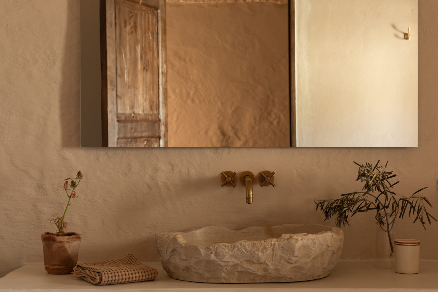 Modern bathroom design featuring a rustic stone sink, brass faucet, and a decorative mirror. A small potted plant and a folded towel add a touch of greenery and softness to the minimalist aesthetic. The warm, earthy tones of the walls and wooden door enhance the cozy atmosphere. | Only on SopranoVillas