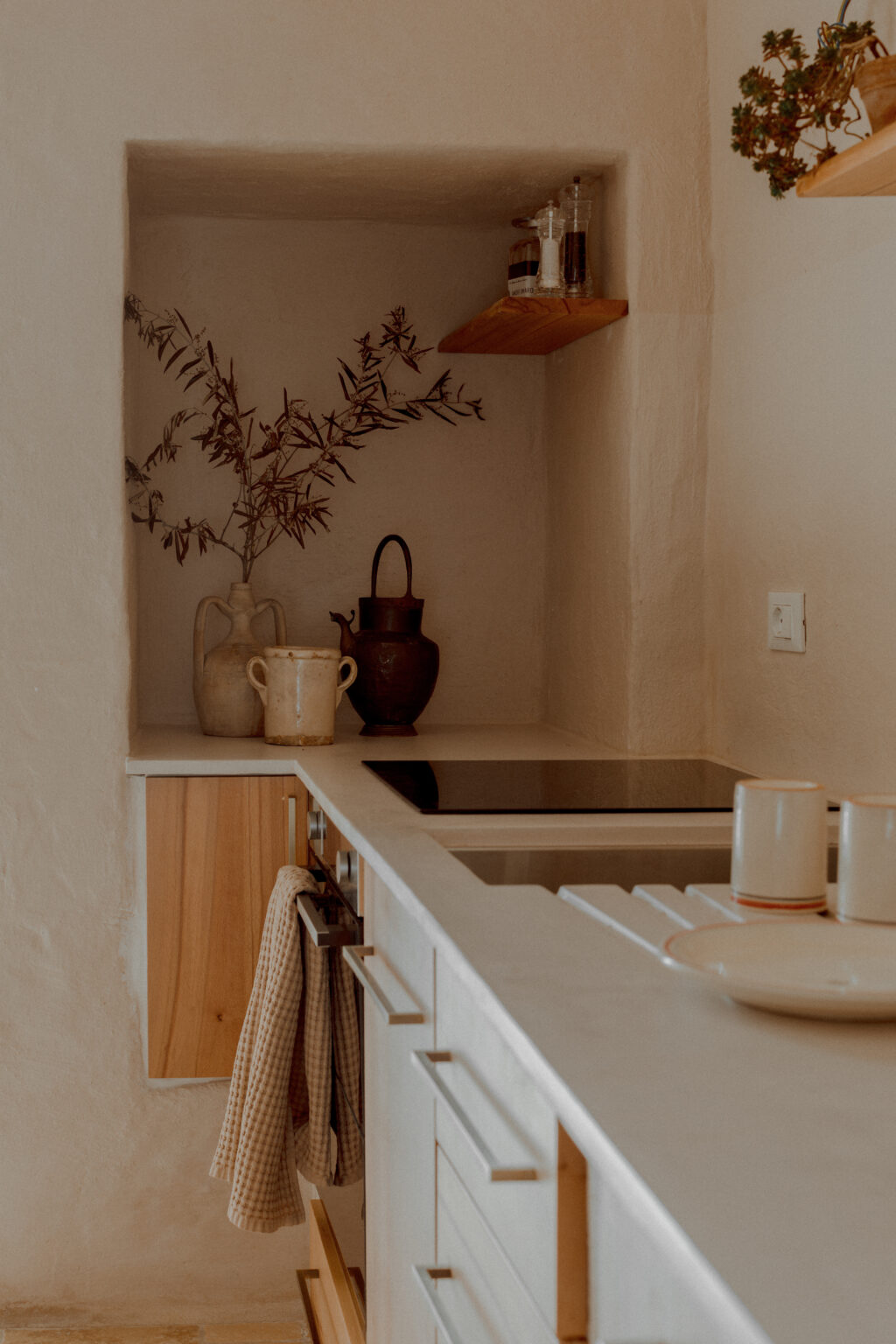 Modern kitchen interior featuring a minimalist design, with a light color palette, wooden accents, and decorative elements like a plant and pottery. The countertop includes an induction cooktop, while a checked towel hangs from a drawer. | Only on SopranoVillas