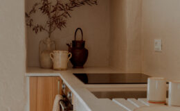 Modern kitchen interior featuring a minimalist design, with a light color palette, wooden accents, and decorative elements like a plant and pottery. The countertop includes an induction cooktop, while a checked towel hangs from a drawer.