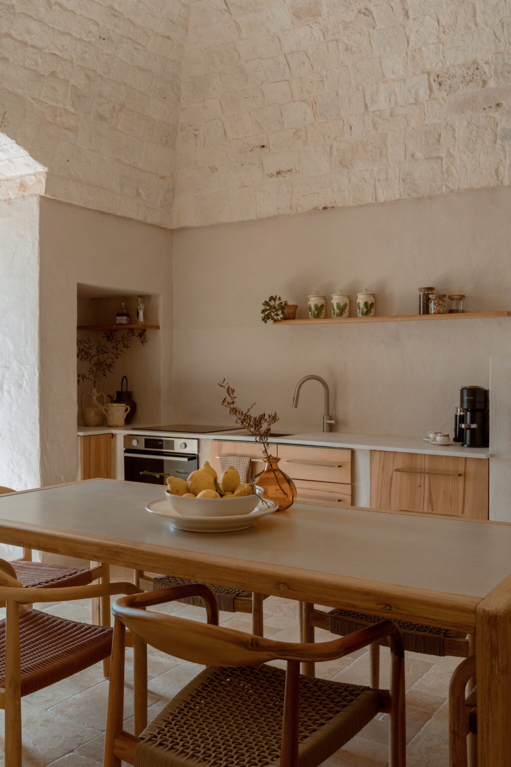 Modern kitchen interior featuring a wooden dining table with a bowl of lemons, stylish chairs, and a minimalist design. The kitchen boasts natural stone walls, a sleek countertop, and wooden cabinetry, creating a warm and inviting atmosphere. | Only on SopranoVillas