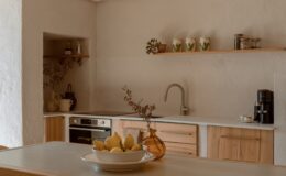 Modern kitchen interior featuring a wooden dining table with a bowl of lemons, stylish chairs, and a minimalist design. The kitchen boasts natural stone walls, a sleek countertop, and wooden cabinetry, creating a warm and inviting atmosphere.