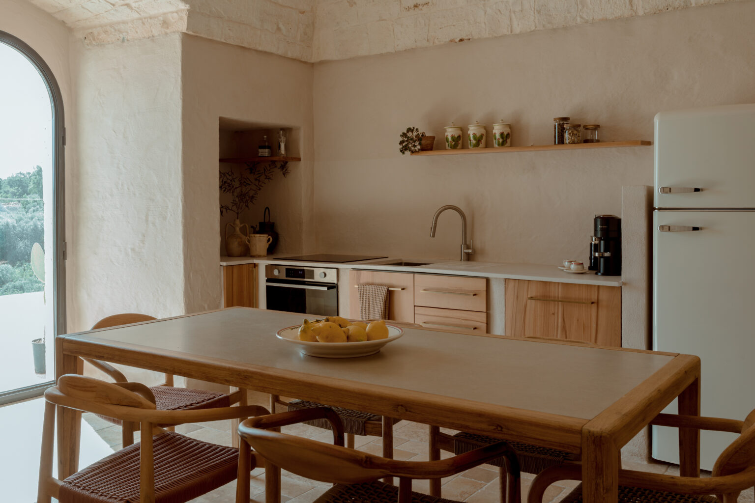 Modern kitchen interior featuring a wooden dining table with a bowl of lemons, minimalist cabinetry, and sleek appliances. Natural light floods the space through a large window, highlighting the earthy color palette and stylish decor elements. | Only on SopranoVillas