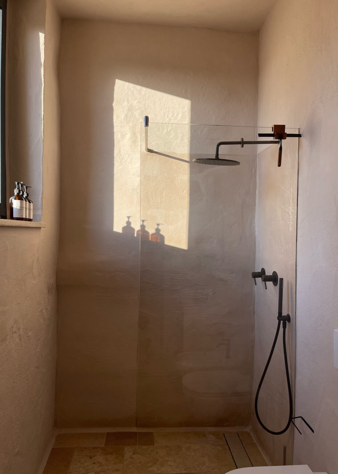 Modern bathroom shower with a glass enclosure, featuring a rainfall showerhead and minimalist decor. Natural light casts shadows on the textured wall, highlighting the sleek design. Bottles of shower products are neatly arranged on the windowsill. | Only on SopranoVillas