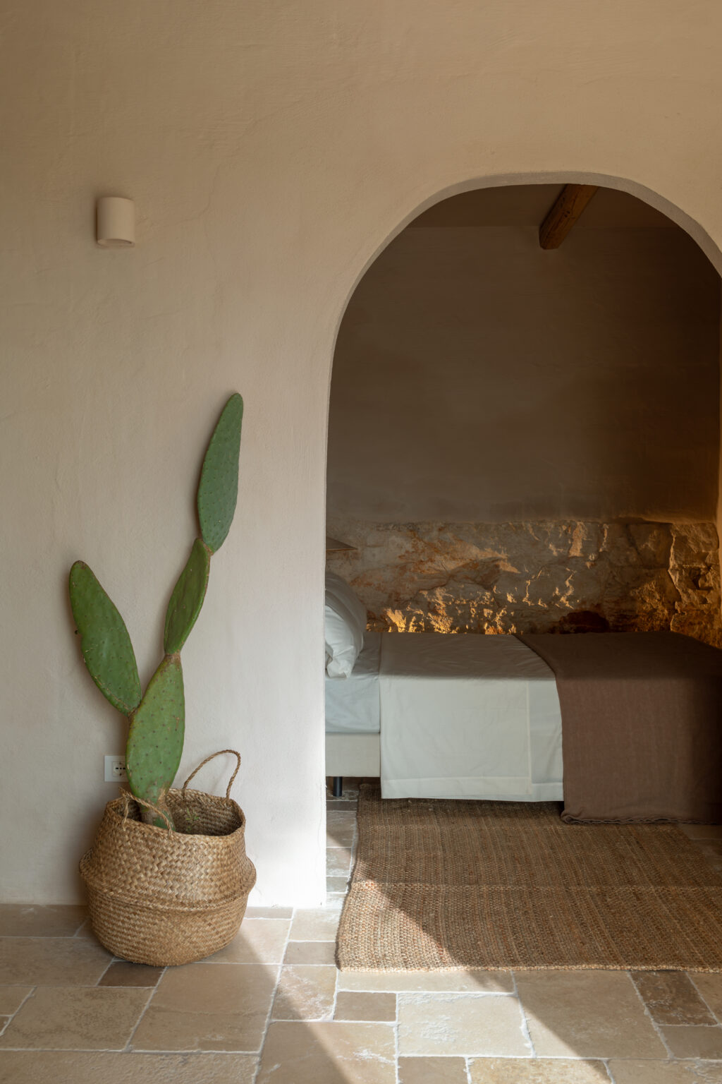Interior view of a minimalist bedroom featuring a large arched doorway, a cactus plant in a woven basket, and natural stone flooring. The room is softly lit, highlighting a bed with white linens and a brown throw. | Only on SopranoVillas
