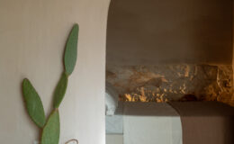Interior view of a minimalist bedroom featuring a large arched doorway, a cactus plant in a woven basket, and natural stone flooring. The room is softly lit, highlighting a bed with white linens and a brown throw.