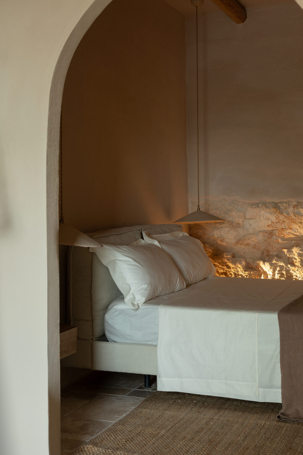 Cozy bedroom featuring a neutral color palette, a plush bed with white pillows and a light beige bedspread, and a warm stone wall illuminated by pendant lighting. The space is designed with minimalist decor, showcasing a natural woven rug and soft textures. | Only on SopranoVillas