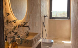 Modern bathroom interior featuring a large circular mirror with backlighting, a stone sink, and stylish fixtures. Natural light streams through a window, illuminating the textured walls and elegant decor, including olive branches and decorative baskets. The space combines minimalist design with a warm, inviting atmosphere.