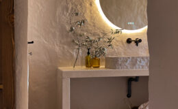 Modern bathroom interior featuring a minimalist design, stone sink, illuminated round mirror, and natural decor elements like olive branches. The warm-toned walls and textured stone flooring create a serene atmosphere.