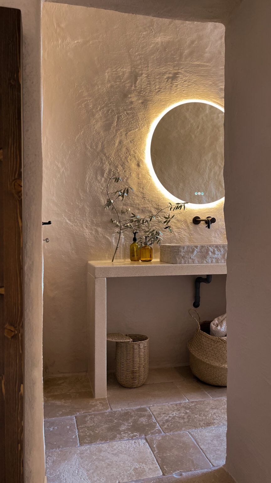 Modern bathroom interior featuring a minimalist design, stone sink, illuminated round mirror, and natural decor elements like olive branches. The warm-toned walls and textured stone flooring create a serene atmosphere. | Only on SopranoVillas