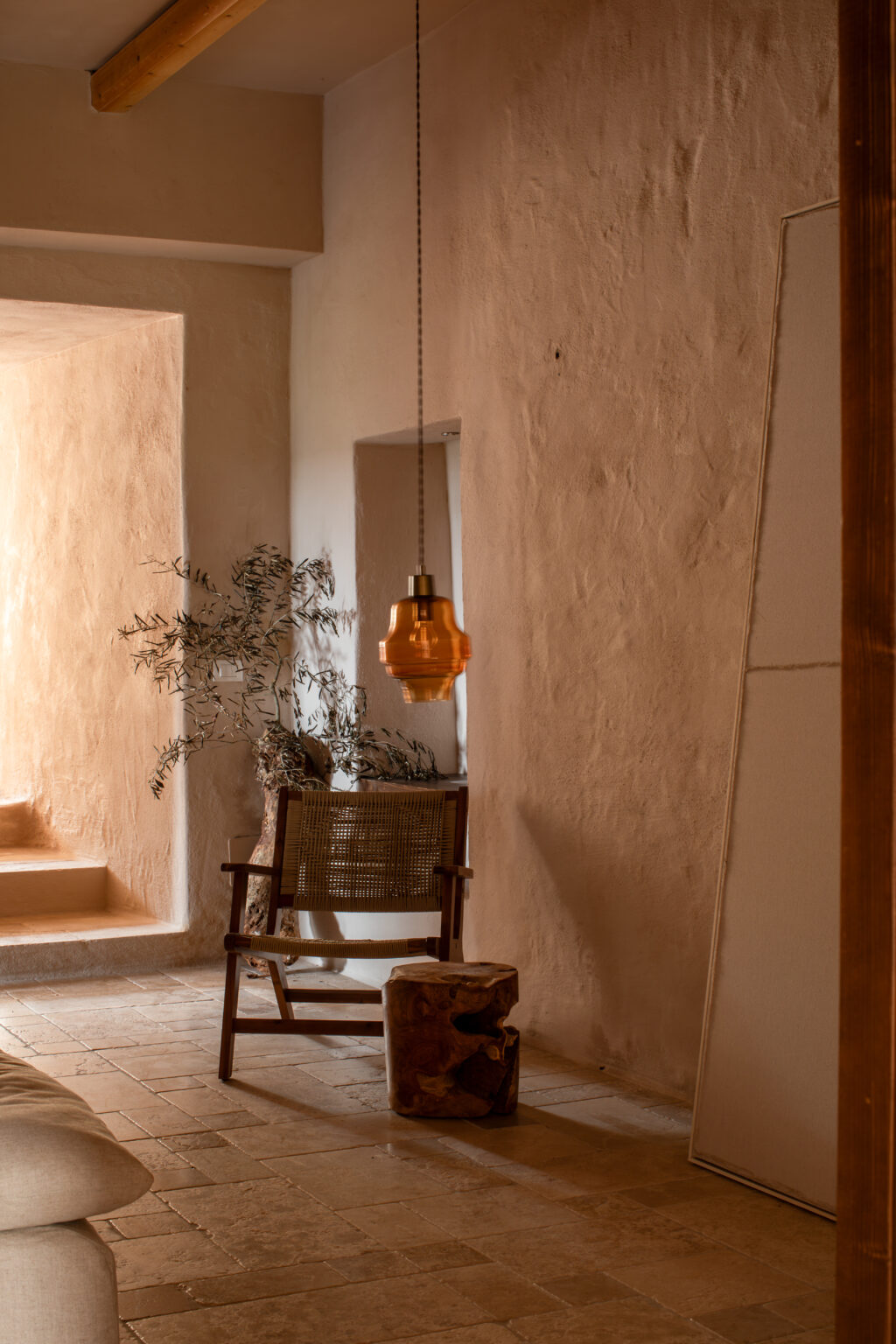 Cozy interior space featuring a wooden chair with woven seating, a rustic stump table, and an amber pendant light hanging from the ceiling, against a backdrop of textured beige walls and natural decor elements like olive branches. | Only on SopranoVillas