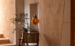 Cozy interior space featuring a wooden chair with woven seating, a rustic stump table, and an amber pendant light hanging from the ceiling, against a backdrop of textured beige walls and natural decor elements like olive branches.