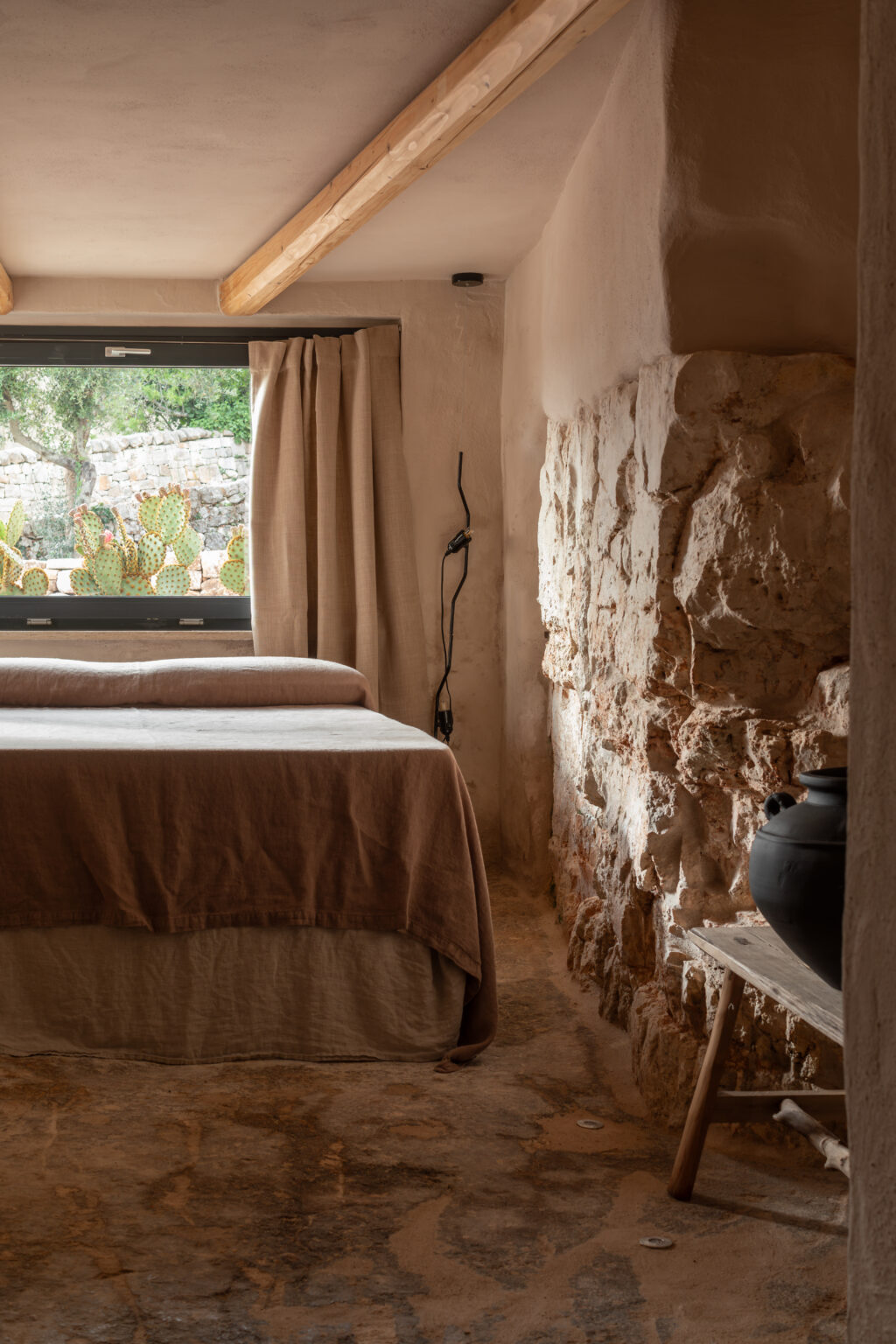 Cozy bedroom interior featuring a minimalist design with a stone wall, wooden ceiling beams, and natural light streaming through a window adorned with beige curtains. The room includes a neatly made bed with neutral-toned bedding and a rustic wooden table displaying a black pot. Cacti visible outside provide a touch of nature. | Only on SopranoVillas