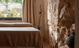Cozy bedroom interior featuring a minimalist design with a stone wall, wooden ceiling beams, and natural light streaming through a window adorned with beige curtains. The room includes a neatly made bed with neutral-toned bedding and a rustic wooden table displaying a black pot. Cacti visible outside provide a touch of nature.