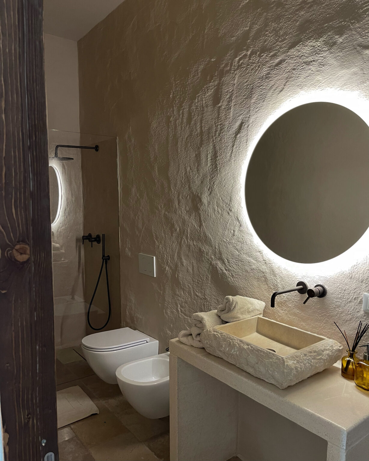 Modern bathroom interior featuring a stone sink, LED circular mirror, and minimalist design. The space includes a wall-mounted faucet, two stylish toilets, and neatly arranged towels, all complemented by earthy tones and natural textures. | Only on SopranoVillas