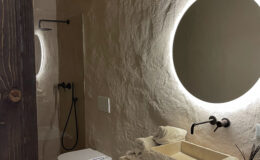 Modern bathroom interior featuring a stone sink, LED circular mirror, and minimalist design. The space includes a wall-mounted faucet, two stylish toilets, and neatly arranged towels, all complemented by earthy tones and natural textures.