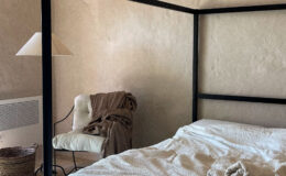 Cozy bedroom featuring a black four-poster bed with a textured cream blanket, neatly arranged towels, and a soft, neutral-toned armchair draped with a blanket. The room has a minimalist aesthetic with warm, natural lighting and a woven basket on the floor.
