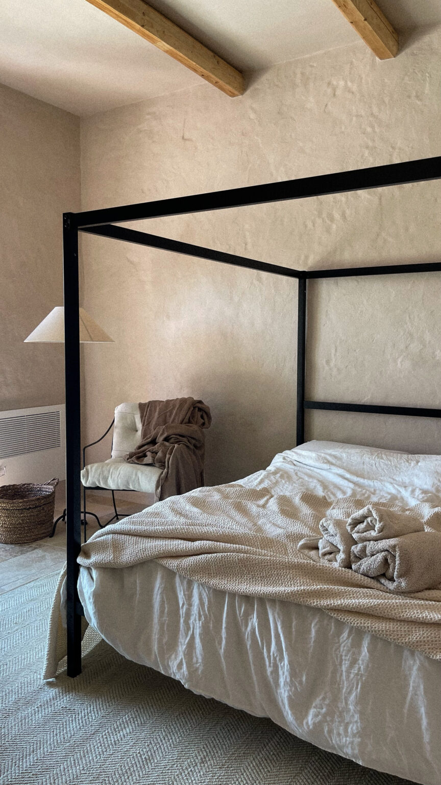 Cozy bedroom featuring a black four-poster bed with a textured cream blanket, neatly arranged towels, and a soft, neutral-toned armchair draped with a blanket. The room has a minimalist aesthetic with warm, natural lighting and a woven basket on the floor. | Only on SopranoVillas