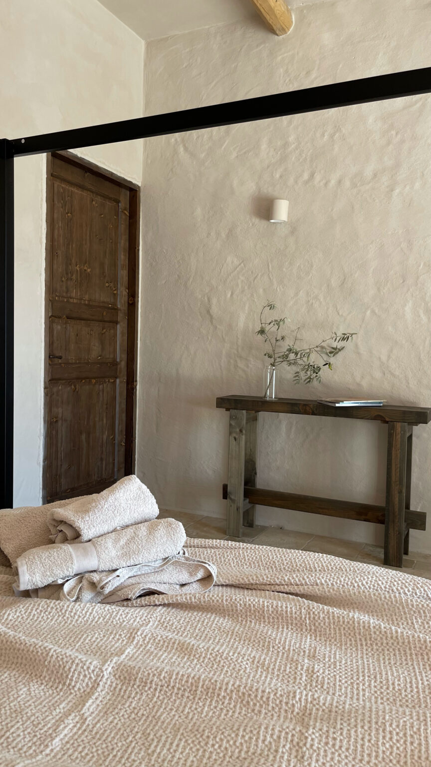 Cozy bedroom interior featuring a neatly made bed with textured bedding and folded towels, a wooden table with a vase containing greenery, and a rustic wooden door in the background. Natural light highlights the warm, earthy tones of the decor. | Only on SopranoVillas