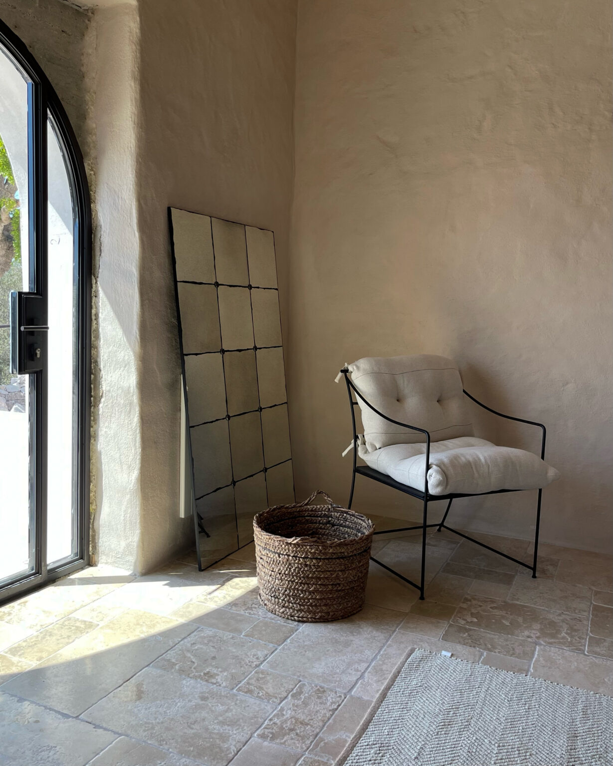 Cozy interior featuring a modern black metal chair with a cushioned seat, a woven basket, and a tall mirror against a textured beige wall. Natural light illuminates the stone floor, creating a warm and inviting atmosphere. | Only on SopranoVillas