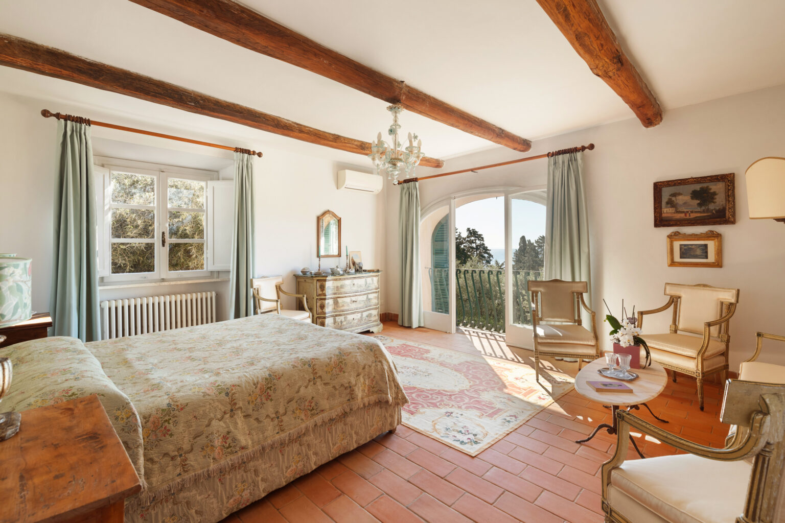 Cozy and elegant bedroom featuring a floral-patterned bedspread, wooden beams, and large windows with scenic views. The room includes antique furniture, a chandelier, and a charming seating area with a small table, creating a warm and inviting atmosphere. | Only on SopranoVillas
