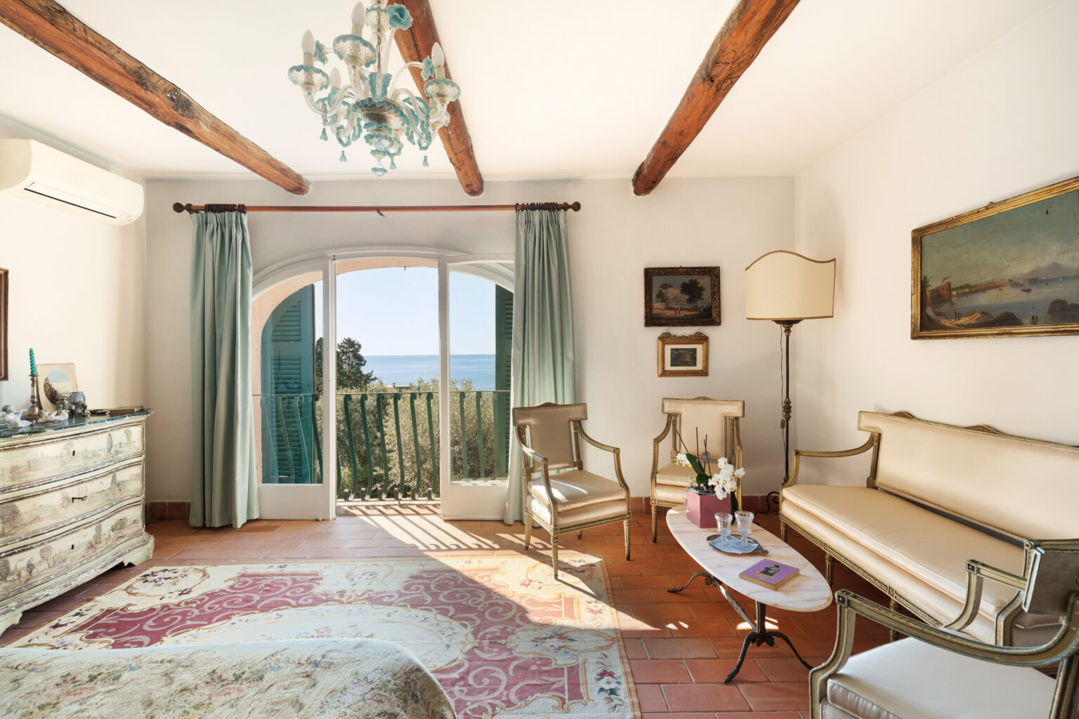 Bright and airy bedroom with a coastal view, featuring a vintage chandelier, wooden beams, and elegant furnishings. The room includes a decorative rug, a chest of drawers, and a cozy seating area with a sofa and chairs, all complemented by natural light streaming through large windows. | Only on SopranoVillas