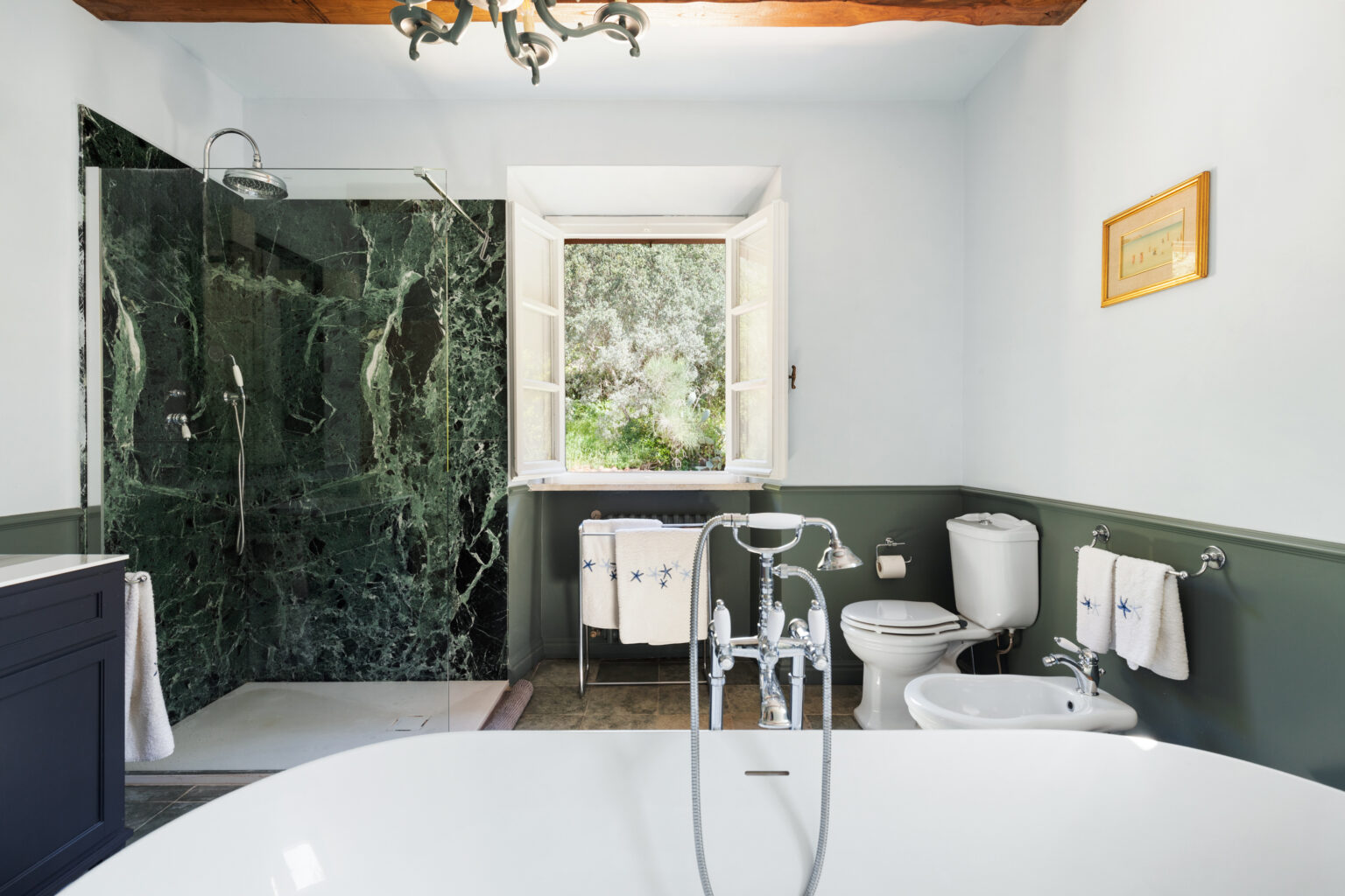 Modern bathroom featuring a freestanding white bathtub, a glass shower with green marble walls, and a window overlooking a lush garden. The space includes a stylish sink, toilet, and decorative towels, accentuated by natural light and elegant fixtures. | Only on SopranoVillas