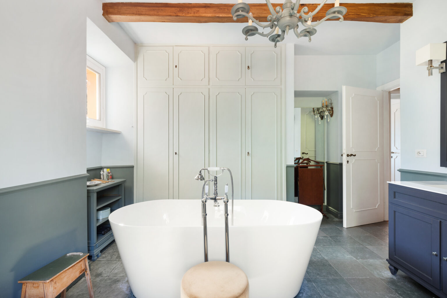 Modern bathroom featuring a freestanding white bathtub, gray stone flooring, and elegant wooden beams. The room includes built-in cabinetry, a stylish chandelier, and a contemporary vanity, creating a serene and sophisticated atmosphere. | Only on SopranoVillas