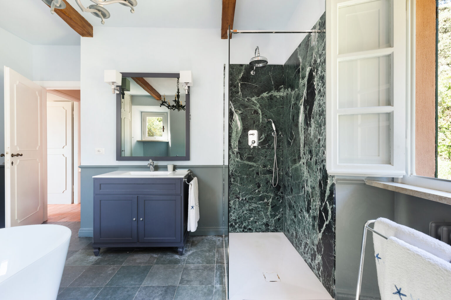 Modern bathroom featuring a sleek design with a glass shower enclosure, green marble walls, a stylish dark cabinet with a white countertop, and a large mirror. Natural light flows through a window, enhancing the space's airy feel. | Only on SopranoVillas