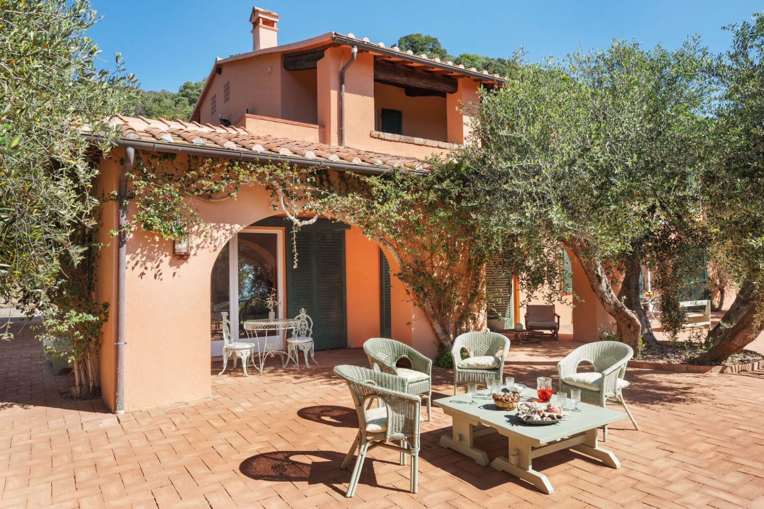 Charming outdoor seating area of a Mediterranean-style villa, featuring a terracotta patio, wicker chairs, and a table adorned with snacks and drinks, surrounded by lush olive trees and vibrant greenery. | Only on SopranoVillas