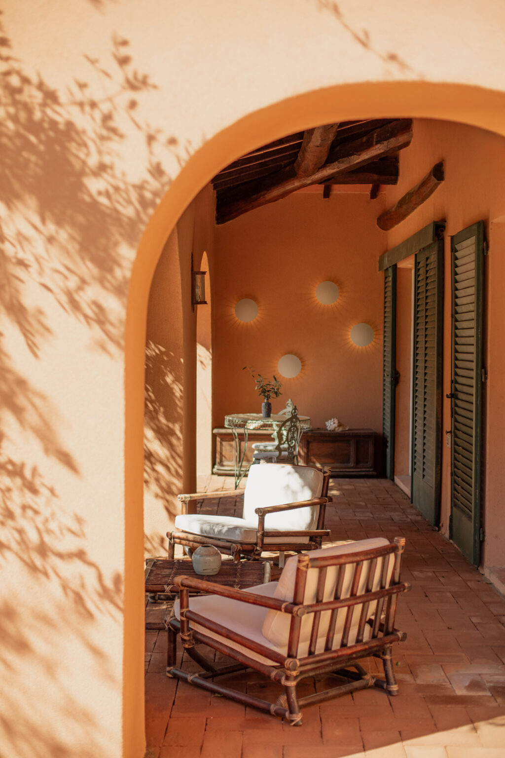 Cozy outdoor seating area with wooden rocking chairs and a small table, framed by an archway. The warm peach-colored walls feature decorative round mirrors and a rustic wooden ceiling. Sunlight casts gentle shadows, creating a serene ambiance in this inviting space. | Only on SopranoVillas