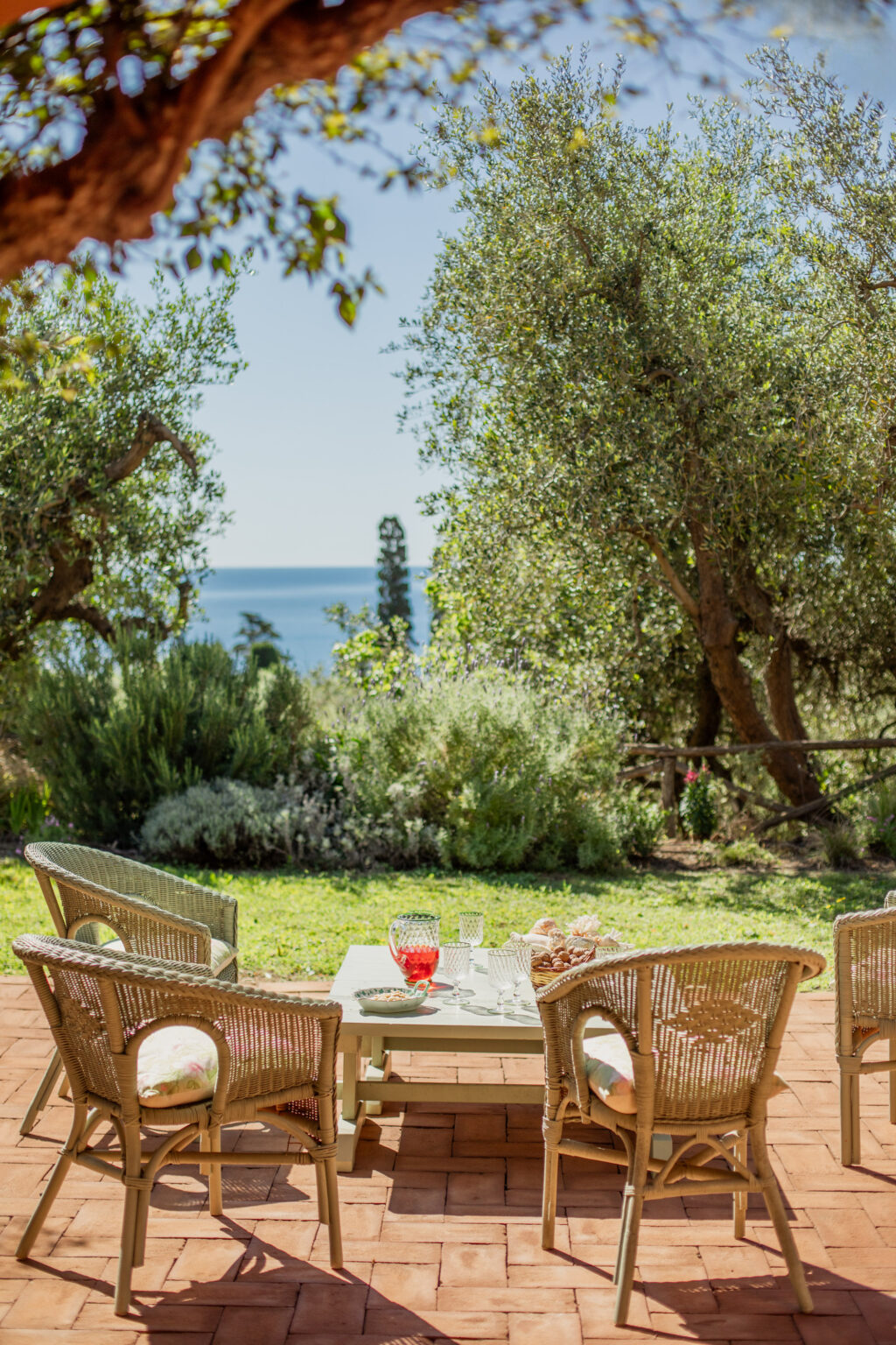 Outdoor seating area with wicker chairs and a table set for refreshments, featuring a scenic view of the ocean and lush greenery in the background. Perfect for relaxation and entertaining in a serene environment. | Only on SopranoVillas