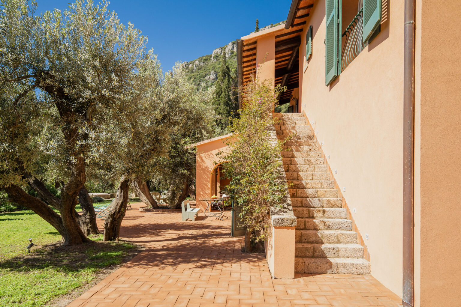 Charming Mediterranean villa exterior featuring a terracotta pathway, olive trees, and a stone staircase leading to a sunny balcony with green shutters against a backdrop of mountains and blue sky. | Only on SopranoVillas