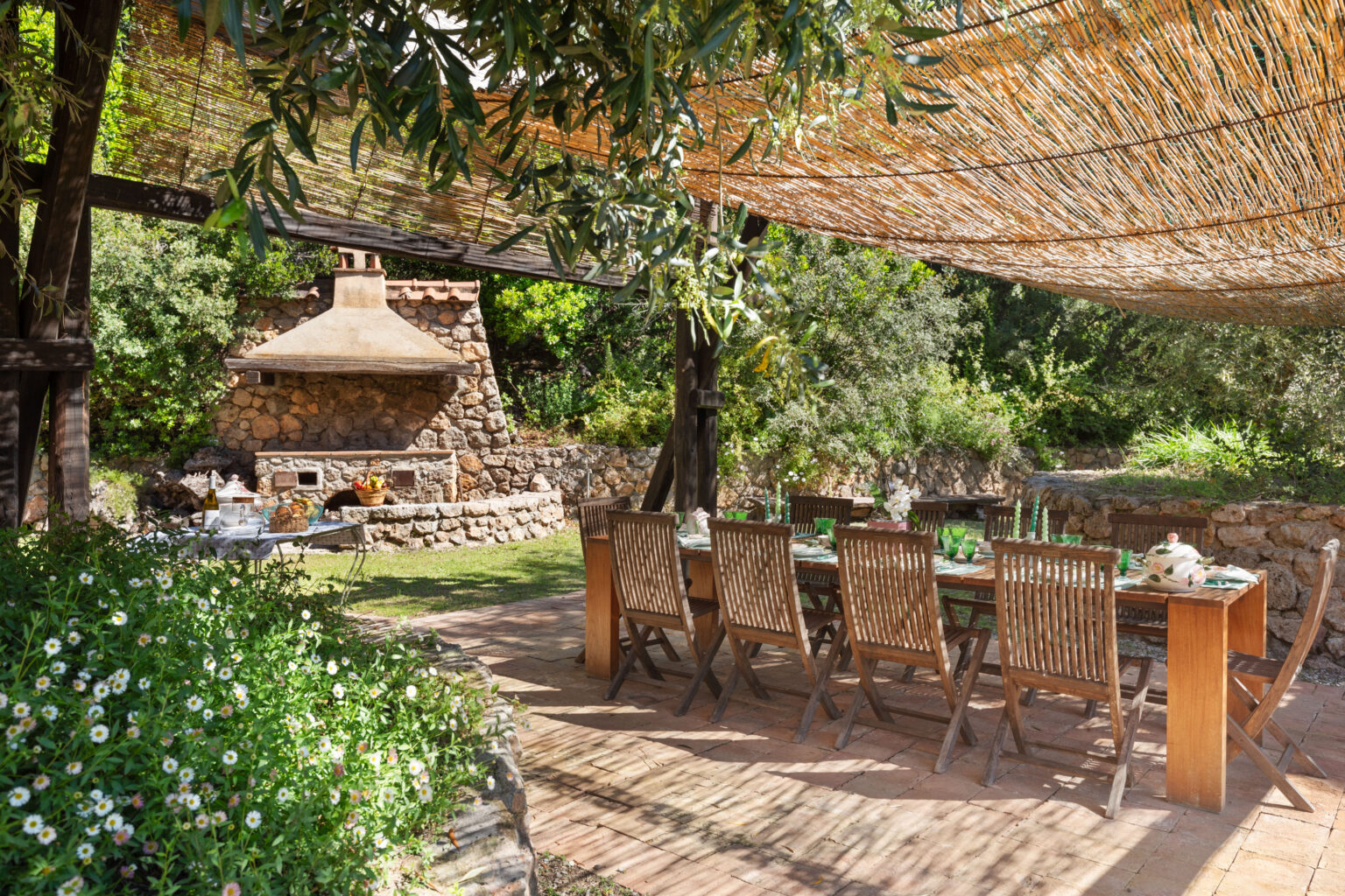 Outdoor dining area featuring a wooden table with green glassware, surrounded by lush greenery and a stone barbecue grill under a shaded pergola. Ideal for summer gatherings and al fresco dining experiences. | Only on SopranoVillas