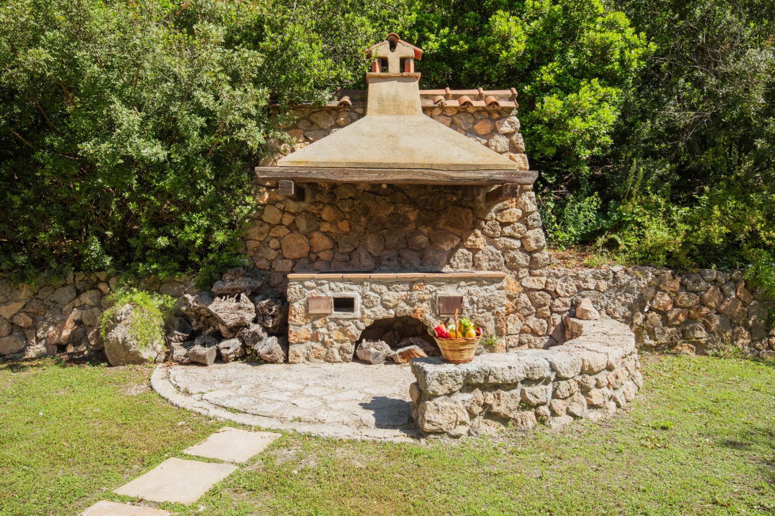 Outdoor stone barbecue grill surrounded by lush greenery, featuring a rustic design with a chimney and a stone seating area, complemented by a basket of fresh fruits in the foreground. Perfect for outdoor cooking and entertaining. | Only on SopranoVillas